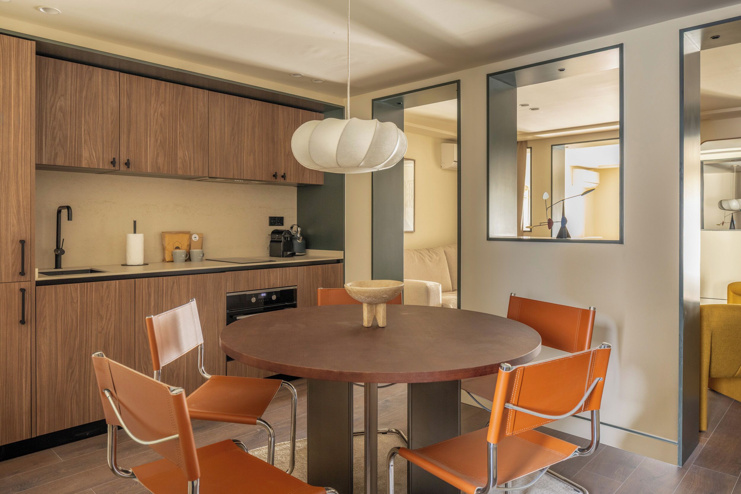 A modern kitchen with wooden cabinets, a round dining table with orange chairs, and a hanging light fixture.