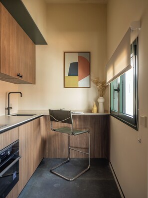 Una cocina moderna con armarios de madera, encimera negra, horno integrado, una silla, un jarrón con flores secas y una ventana con persianas.