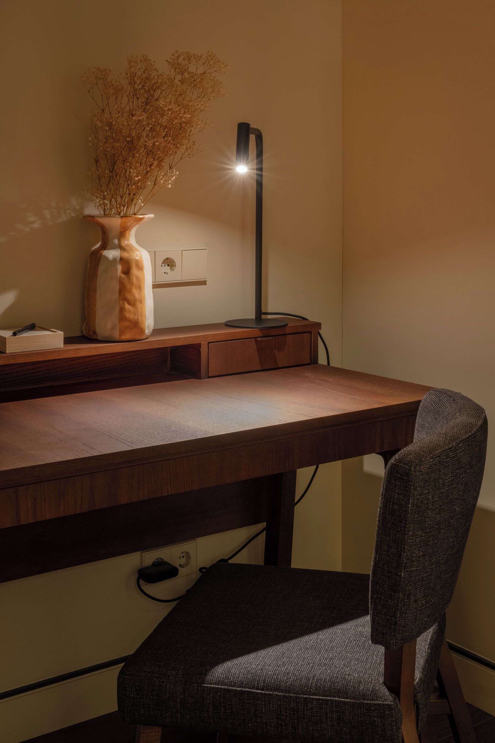 Un escritorio de madera con un jarrón de flores secas, una lámpara y una silla.