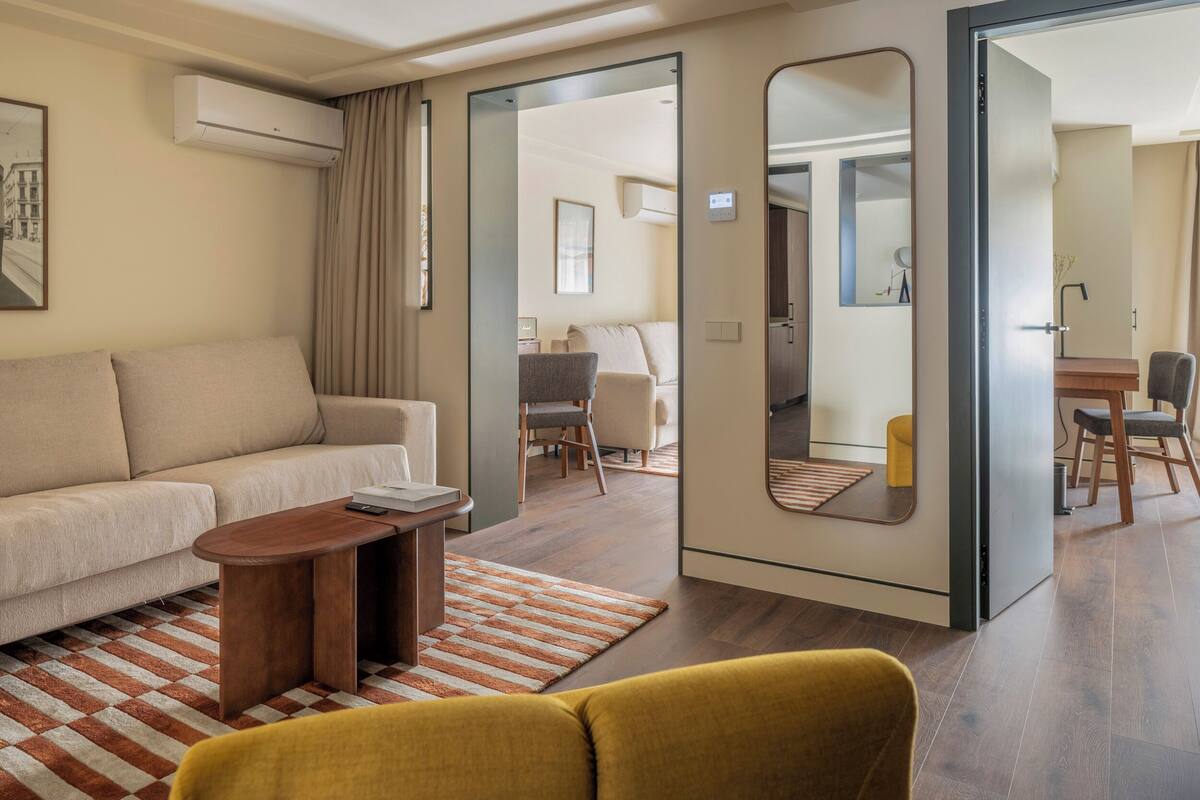 Moderni hotellihuone, jossa on sohva, pieni p öytä, peili ja ruokailutila tuoleineen.