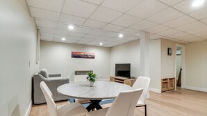 Een moderne woonkamer met een ronde tafel, witte stoelen, een grijze bank, een flatscreen televisie en een schilderij aan de muur.