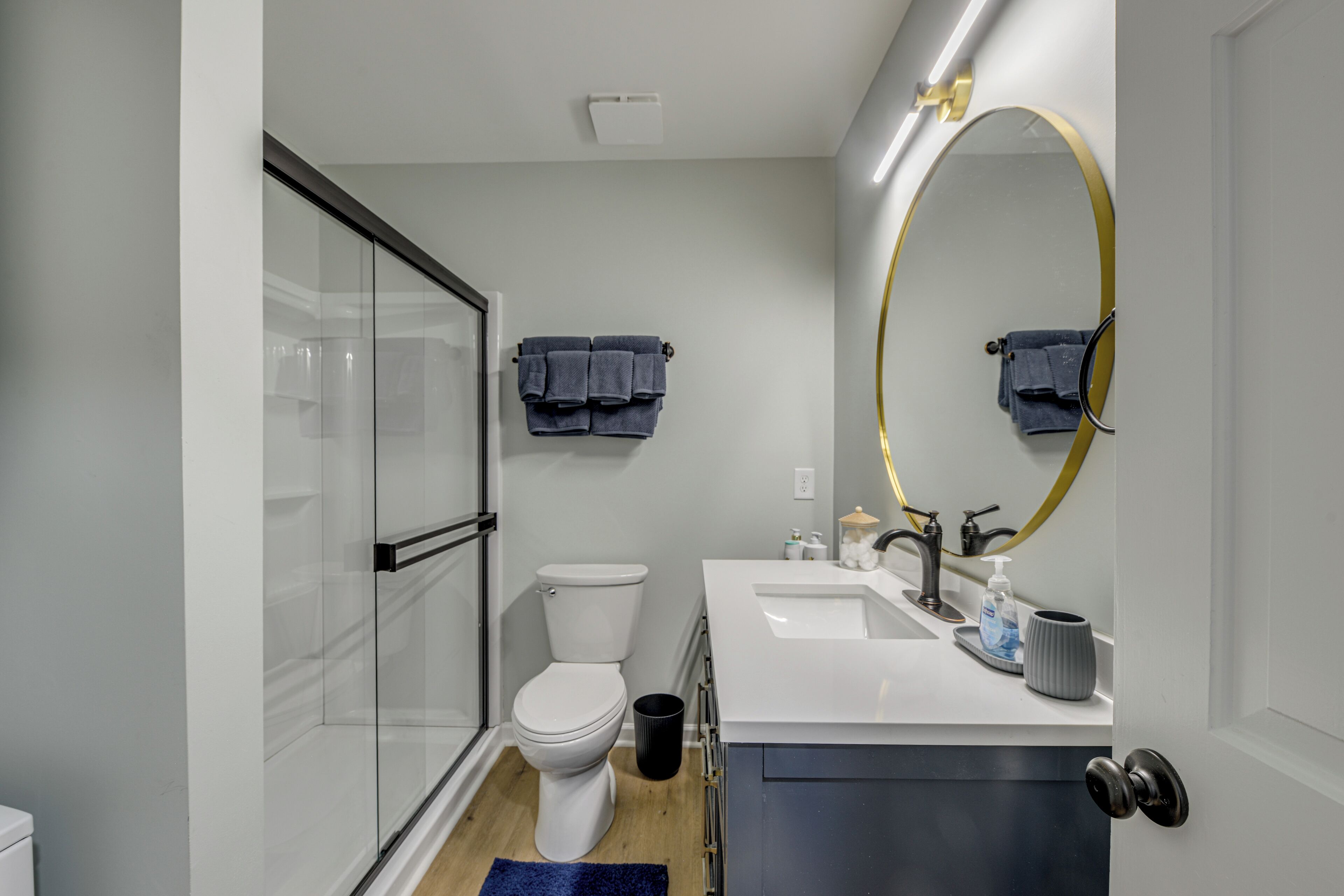 A modern bathroom with a glass shower enclosure, a white toilet, a sink with a gold-framed mirror, and a blue rug.