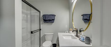 A modern bathroom with a glass shower enclosure, a white toilet, a sink with a gold-framed mirror, and a blue rug.