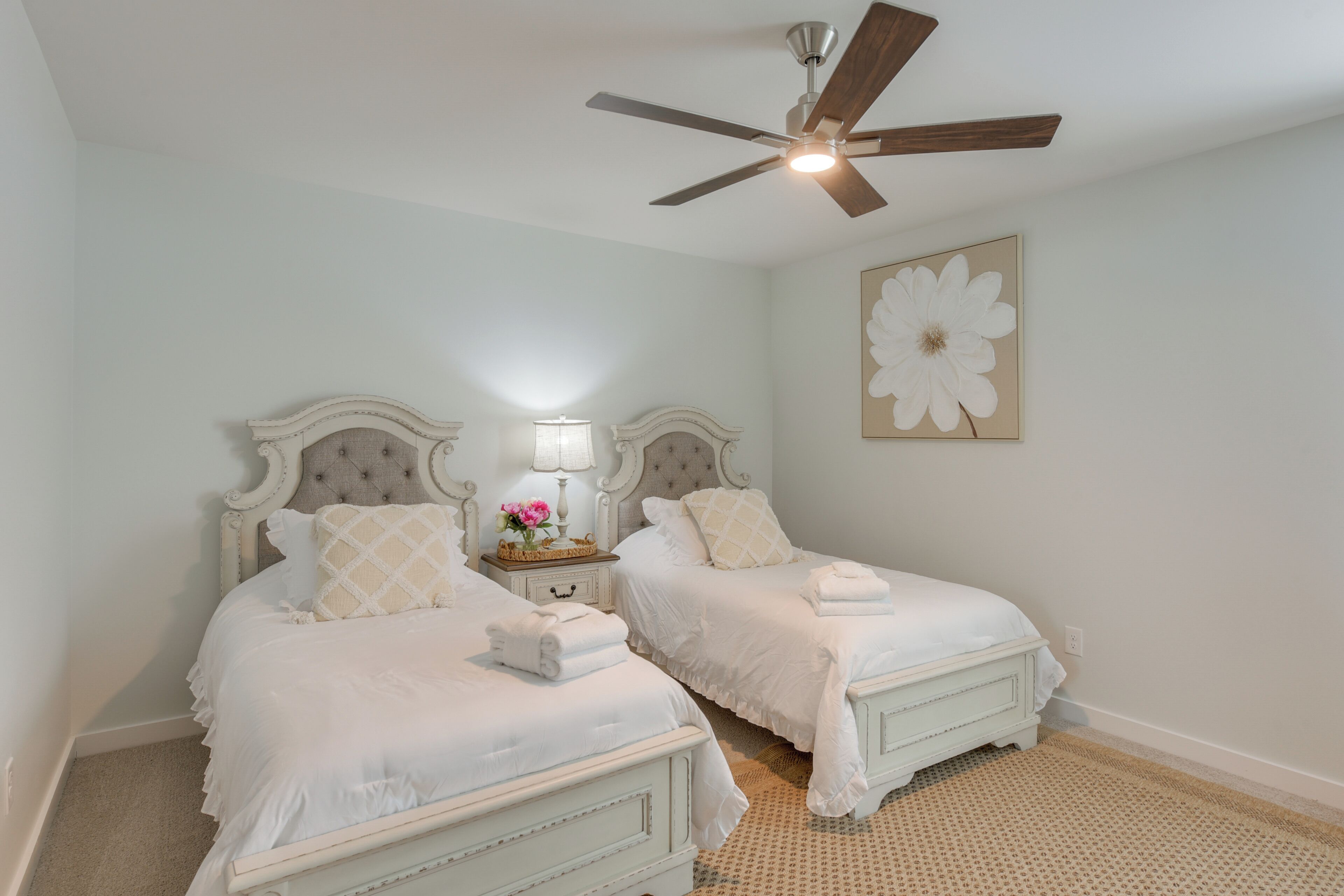 A bedroom with two white beds, a ceiling fan, a nightstand with a lamp, and a wall-mounted flower painting.