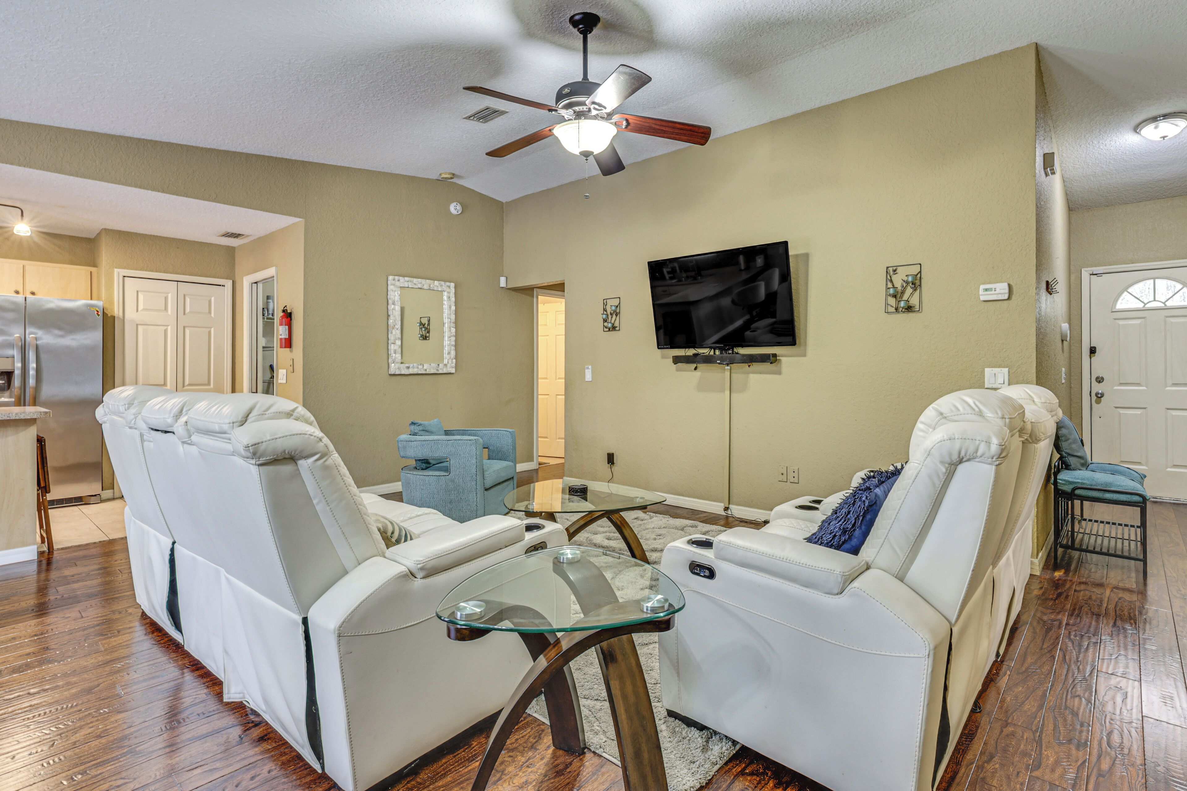 A living room with two white leather sofas, a glass coffee table, a ceiling fan, a flat-screen TV, and a kitchen area in the background.