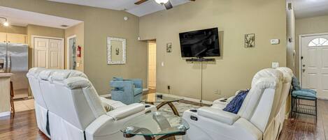 A living room with two white leather sofas, a glass coffee table, a ceiling fan, a flat-screen TV, and a kitchen area in the background.