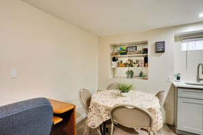 Un pequeño comedor con una mesa cubierta con mantel, rodeada de sillas. Encima de la mesa hay una estantería con varios objetos y un espejo.
