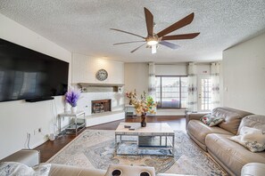 A living room with a fireplace, a ceiling fan, a flat-screen TV mounted on the wall, and a sofa. - Walk to Lake! Home w/ Pool Near At&t Stadium (Rowlett)
