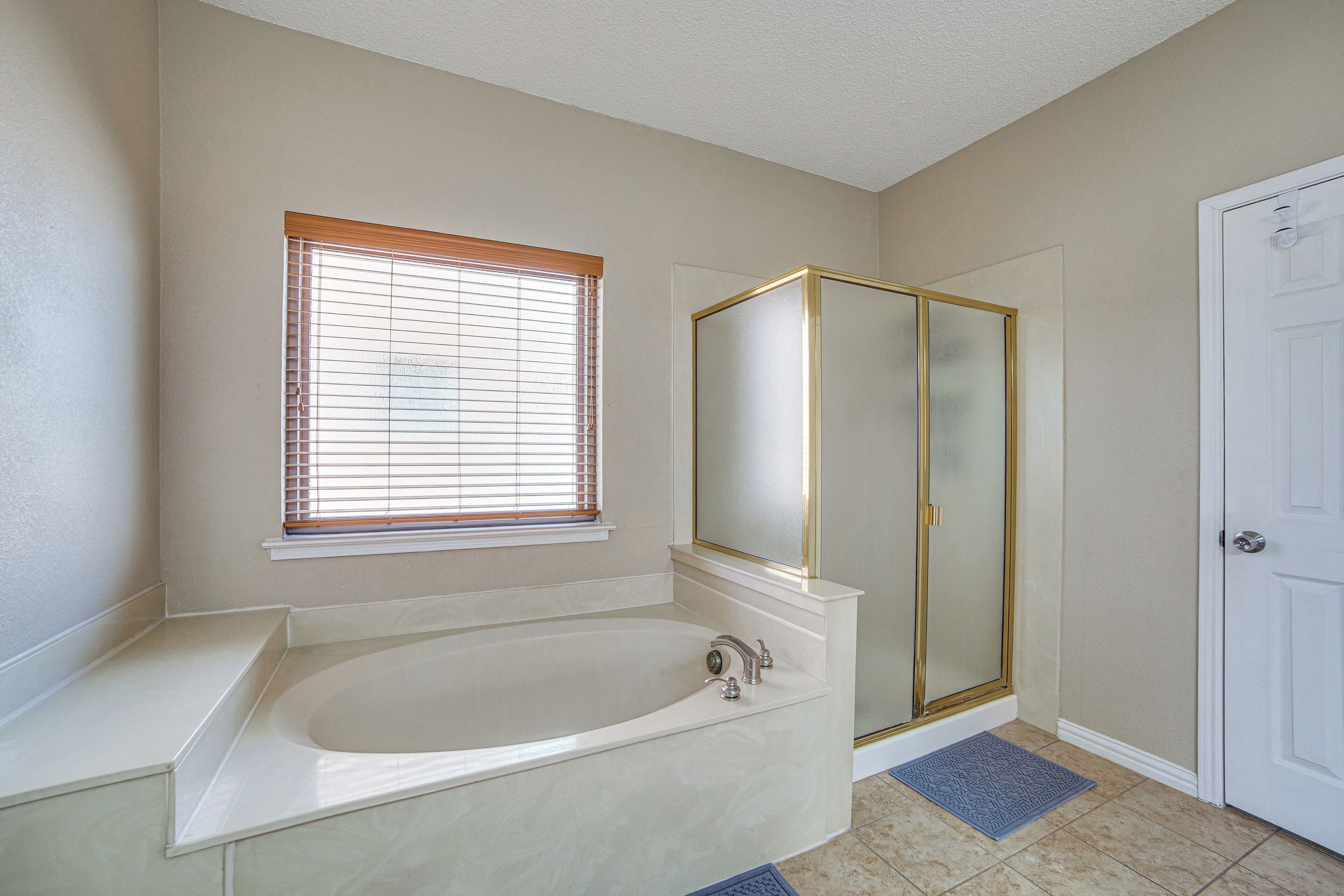 A bathroom with a bathtub, a walk-in shower, a window with blinds, and a door.