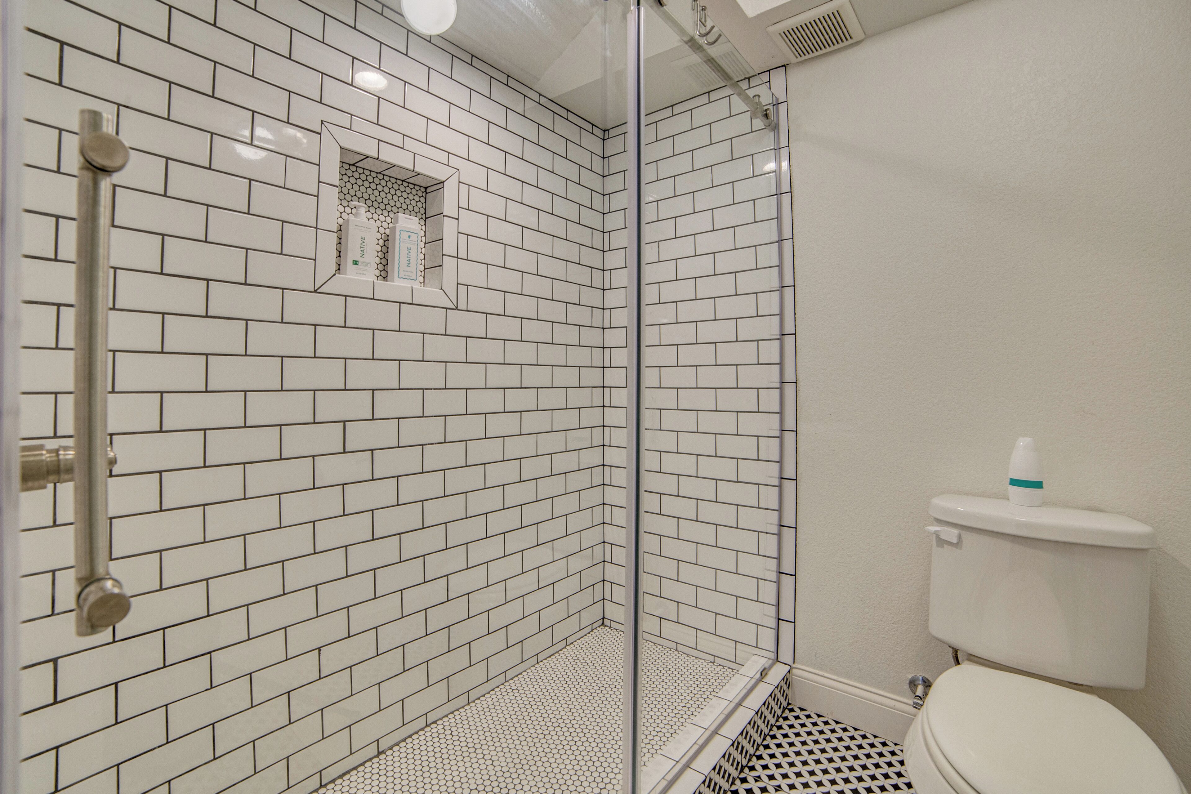 Et badeværelse med hvide, rektangulære fliser, et glasbadeværelsesafskærmning og et toilet.