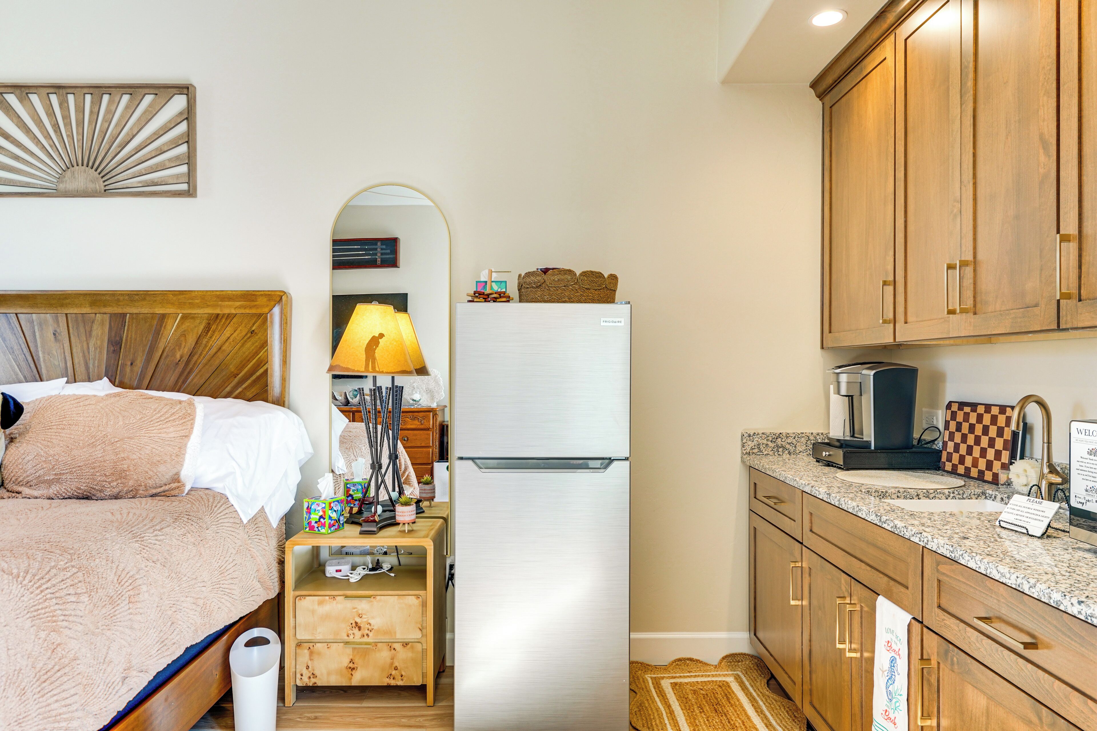 A bedroom with a bed, a nightstand with a lamp, a refrigerator, and a kitchen area with wooden cabinets and a countertop.