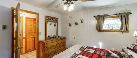 A bedroom with a bed, a wooden dresser, a ceiling fan, a window with curtains, and a door leading to another room.
