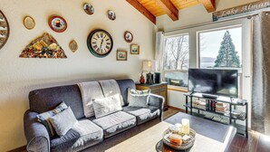 Een woonkamer met een bank, een salontafel en een televisie op een standaard. Er zijn wanddecoraties en een raam met uitzicht op bomen.