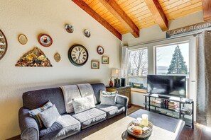 A cozy living room with a sectional sofa, a flat-screen TV, and a large window offering a view of a mountain and a tree. - Private Boat Slip & Views: Lake Almanor Getaway! (Westwood)