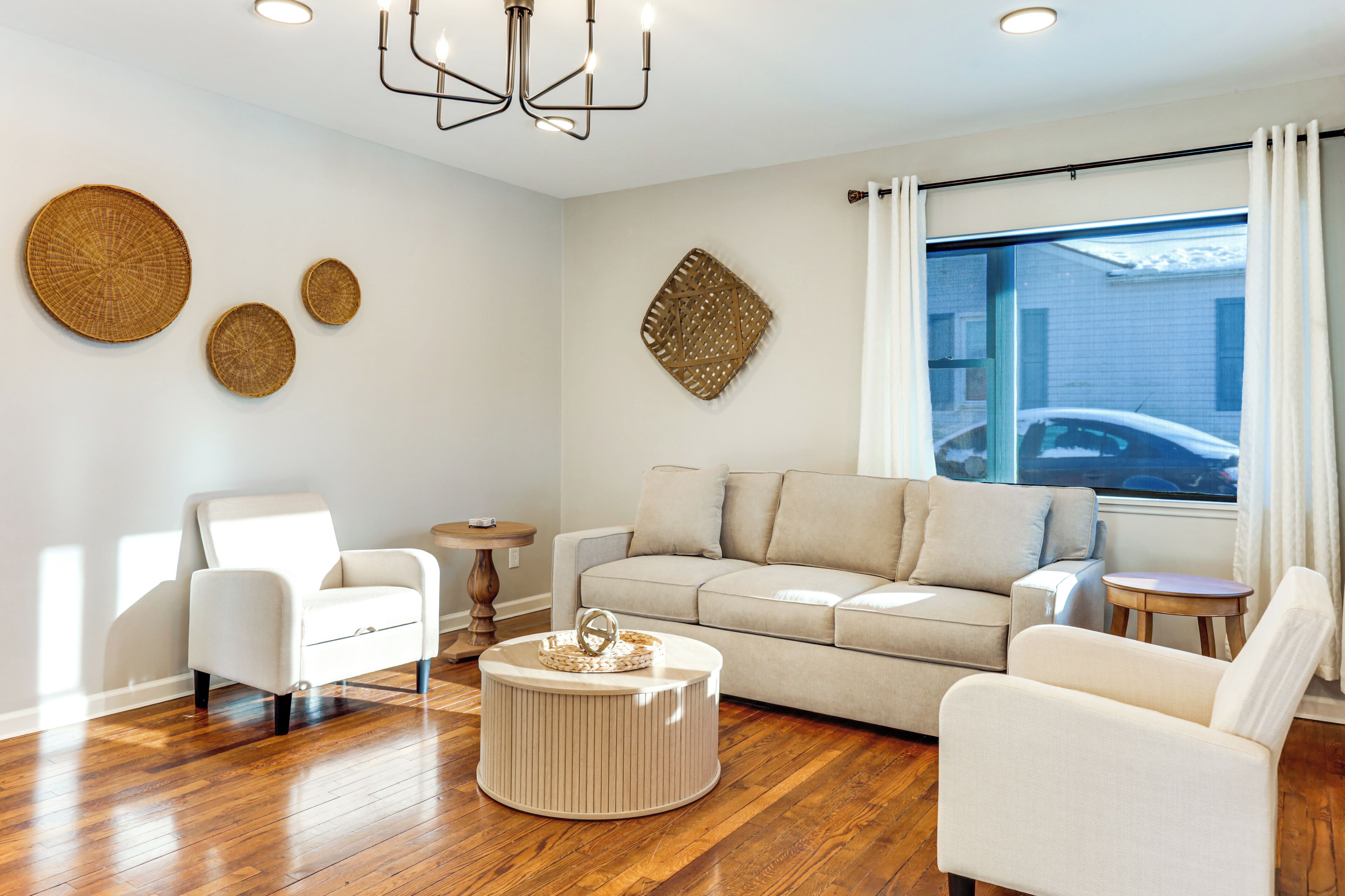 A modern living room with a beige sofa, two white armchairs, a round coffee table, and a chandelier.