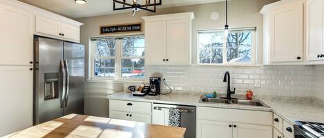 A modern kitchen with white cabinets, a stainless steel refrigerator, a wooden island, and a window with a view of trees.