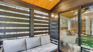 A wooden deck with a sofa and a glass door leading to a bedroom. - Fire Pit, Deck! Modern Tiny Home in Piney Woods (Linden)