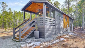 A cabin with a wooden staircase leading to a covered porch, surrounded by trees. - Fire Pit, Deck! Modern Tiny Home in Piney Woods (Linden)