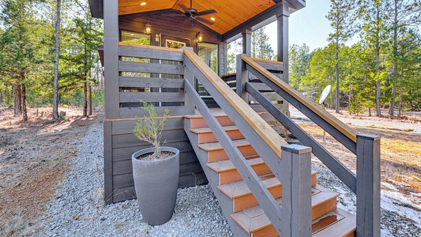 A wooden staircase leading to a covered area with a ceiling fan and lights, surrounded by trees. - Fire Pit, Deck! Modern Tiny Home in Piney Woods (Linden)