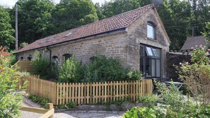 A stone building with a tiled roof, a wooden fence, and a garden with various plants. - Horse Gin (Lydney)