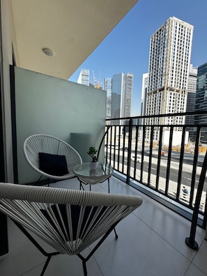 A balcony with a wicker chair, a small table with a plant, and a view of tall buildings. - Stylish 1BR Lake View Retreat  (Dubai)