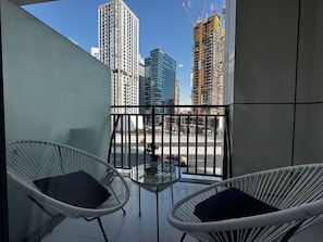 A balcony with two wicker chairs and a small table, overlooking a cityscape with tall buildings and construction cranes. - Stylish 1BR Lake View Retreat  (Dubai)