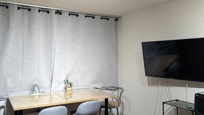 A room with a wooden table, two grey chairs, a white curtain, a flat-screen TV mounted on the wall, and a small shelf with a TV and some items. - Sunny studio for 4 with balcony, lake view, and WiFi (Besse-et-Saint-Anastaise-Super Besse)