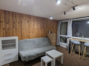 A room with a wooden wall, a grey sofa, a white desk with two chairs, and a window with curtains. - Sunny studio for 4 with balcony, lake view, and WiFi (Besse-et-Saint-Anastaise-Super Besse)