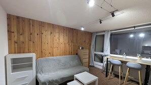 A compact room with a wooden wall, a grey sofa, a small white table, a wooden desk with two chairs, and a window with curtains. - Sunny studio for 4 with balcony, lake view, and WiFi (Besse-et-Saint-Anastaise-Super Besse)