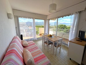 A room with a sofa, a dining table with chairs, a TV, and large windows with a view of greenery and mountains. - 4 Person Apartment with Parking, Sea and Mountain Views (Argelès-sur-Mer)
