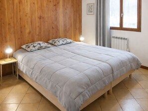 Quarto com cabeceira de madeira, cama com colcha cinza, duas luminárias de cabeceira e janela com cortinas.