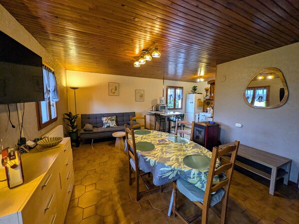 A cozy room with a wooden ceiling, a dining table with chairs, a sofa, a kitchen area, and a mirror on the wall.