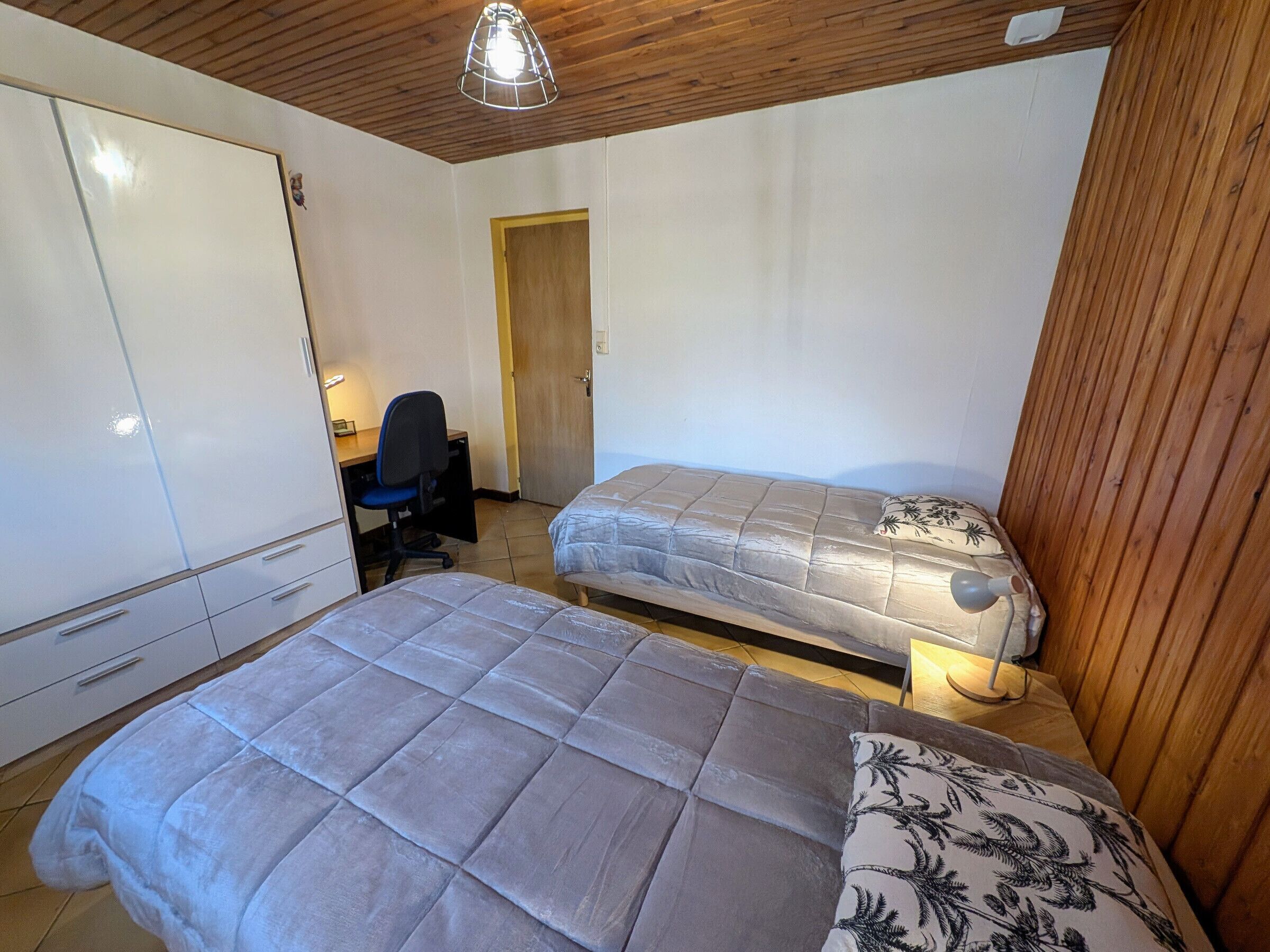 A small, well-lit room with two beds, a desk with a chair, and a wardrobe.