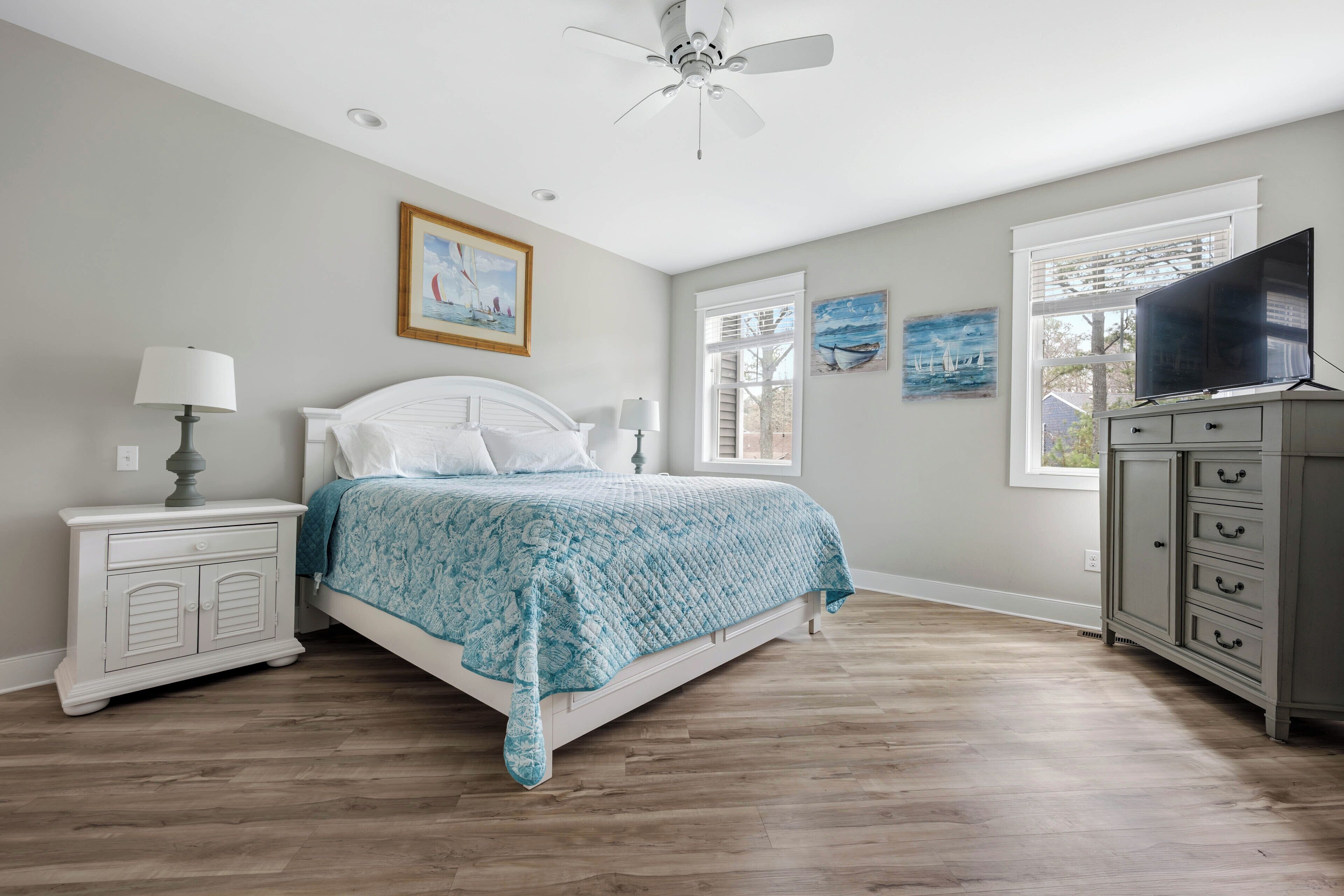 Une chambre à coucher comprenant un lit, une table de chevet, une commode, un ventilateur de plafond et des œuvres d’art accrochées aux murs.