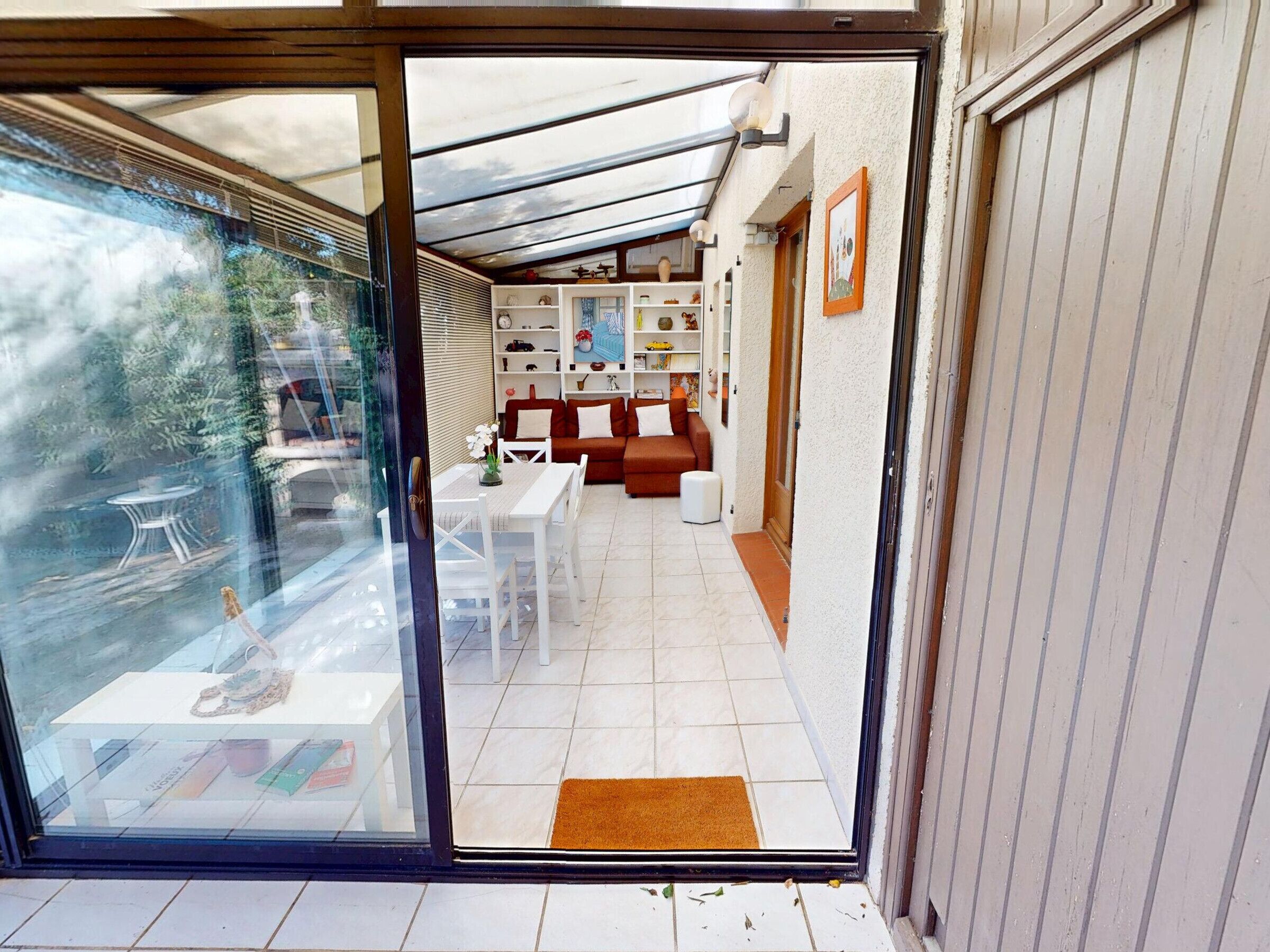 Un salon moderne doté d’une porte vitrée donnant sur un balcon, d’un sol carrelé blanc, d’un canapé marron et d’une table à manger avec des chaises.