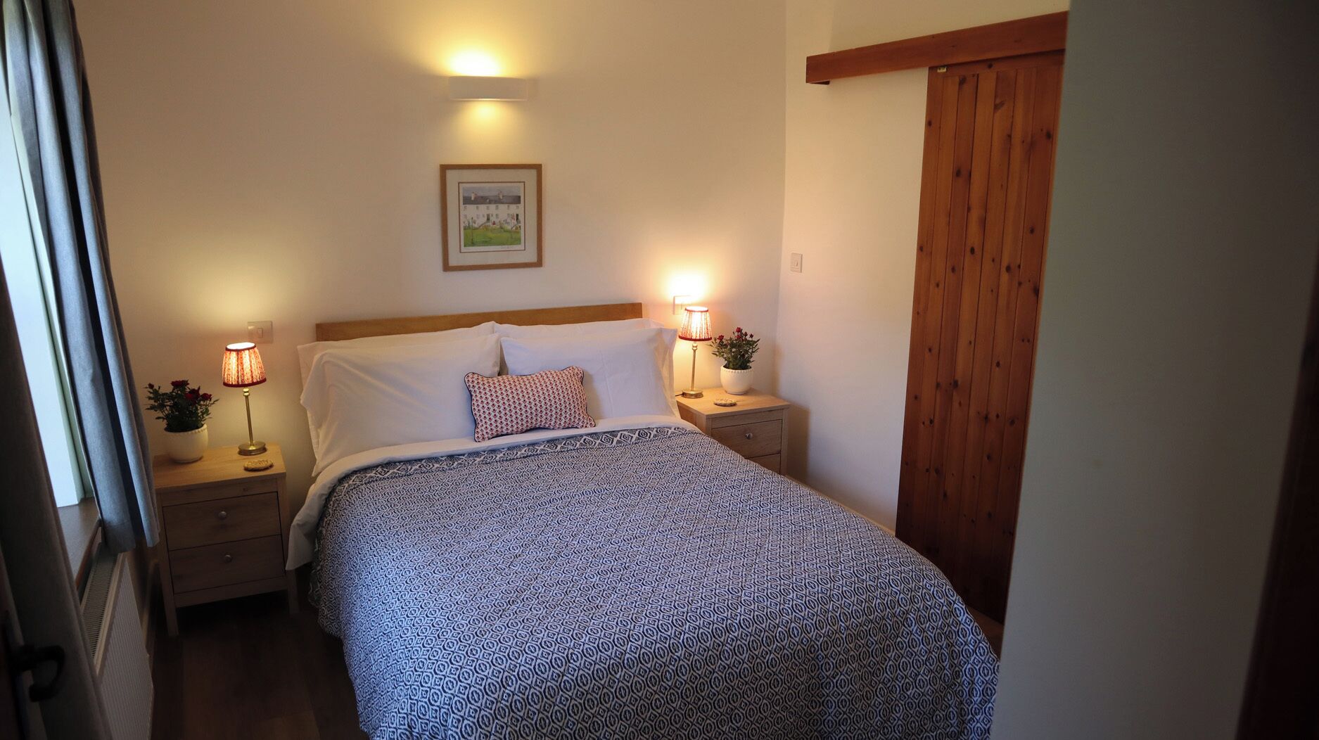 Un dormitorio con una cama, dos mesitas de noche con lámparas, un cuadro en la pared y una puerta de madera.