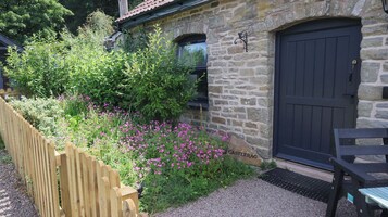 A stone building with a dark door, a wooden fence, and a garden with pink flowers.
