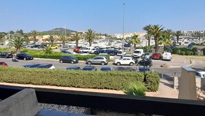 Un puerto deportivo con coches estacionados, barcos, palmeras y edificios al fondo.