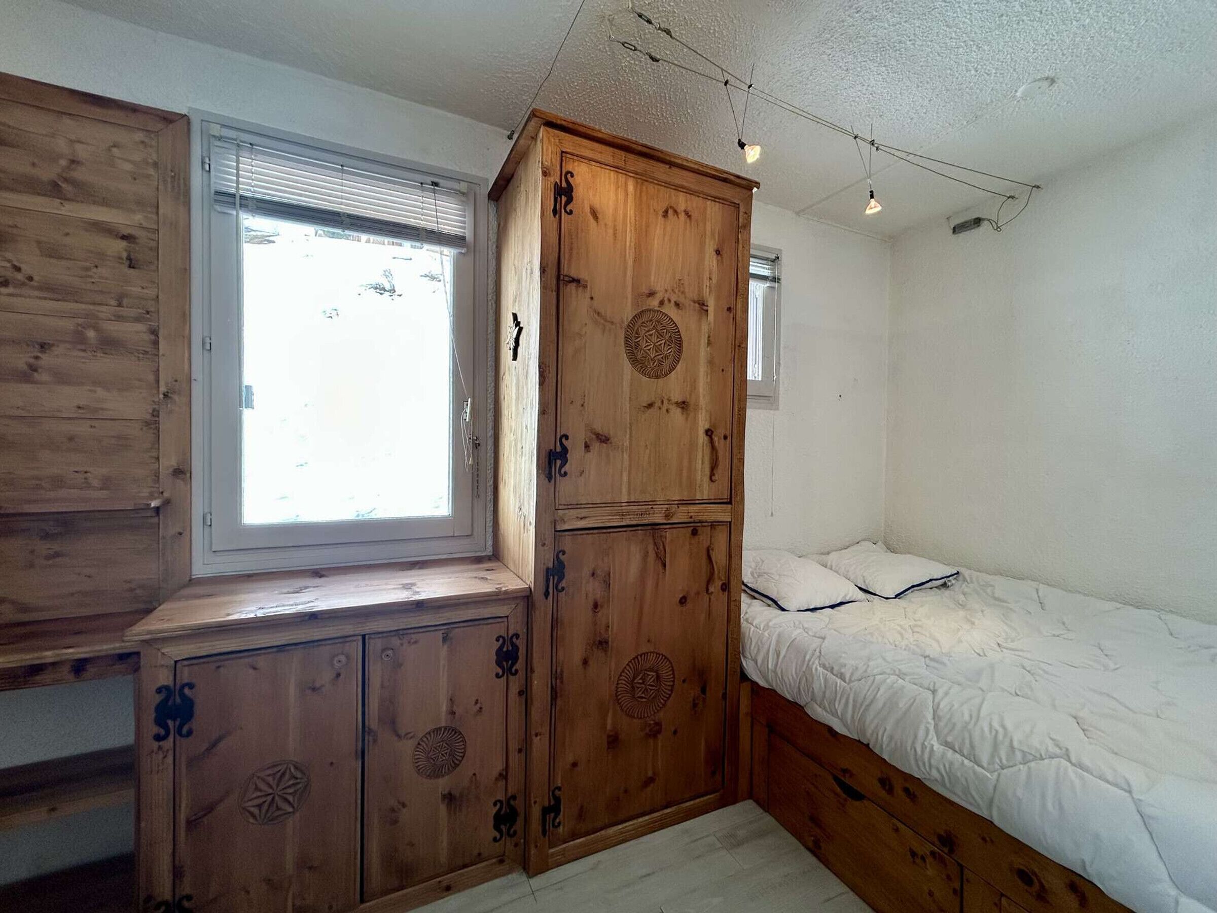 A bedroom with a wooden wardrobe, a window, and a bed.