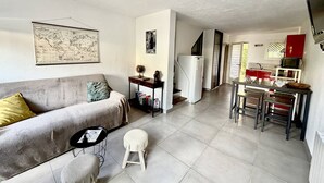 Un espace de vie compact comprenant un canapé, un petit tabouret, une table à manger avec des chaises, un coin cuisine avec un réfrigérateur et une carte accrochée au mur.