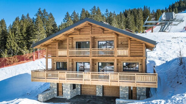 Ein mehrstöckiges Holzchalet mit Balkonen, gelegen an einem verschneiten Hang mit Wald im Hintergrund.