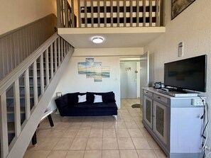 A living room with a staircase, a black sofa, a flat-screen TV, and a cabinet with electronic devices. - Cozy Loft-Style T2 with Pool & Parking, near Argelès-sur-Mer Beach (Argelès-sur-Mer)