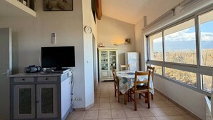 Uma sala de jantar com mesa e cadeiras, uma televisão sobre um móvel, e vista através de amplas janelas.
