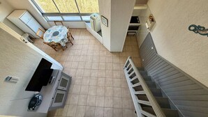 Quarto amplo, com pé direito alto, mesa de jantar e cadeiras, cozinha compacta e vista através de uma grande janela.
