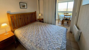 Quarto com cama de madeira, mesa de cabeceira, luminária, janela com cortinas e vista para uma mesa e cadeiras ao ar livre.