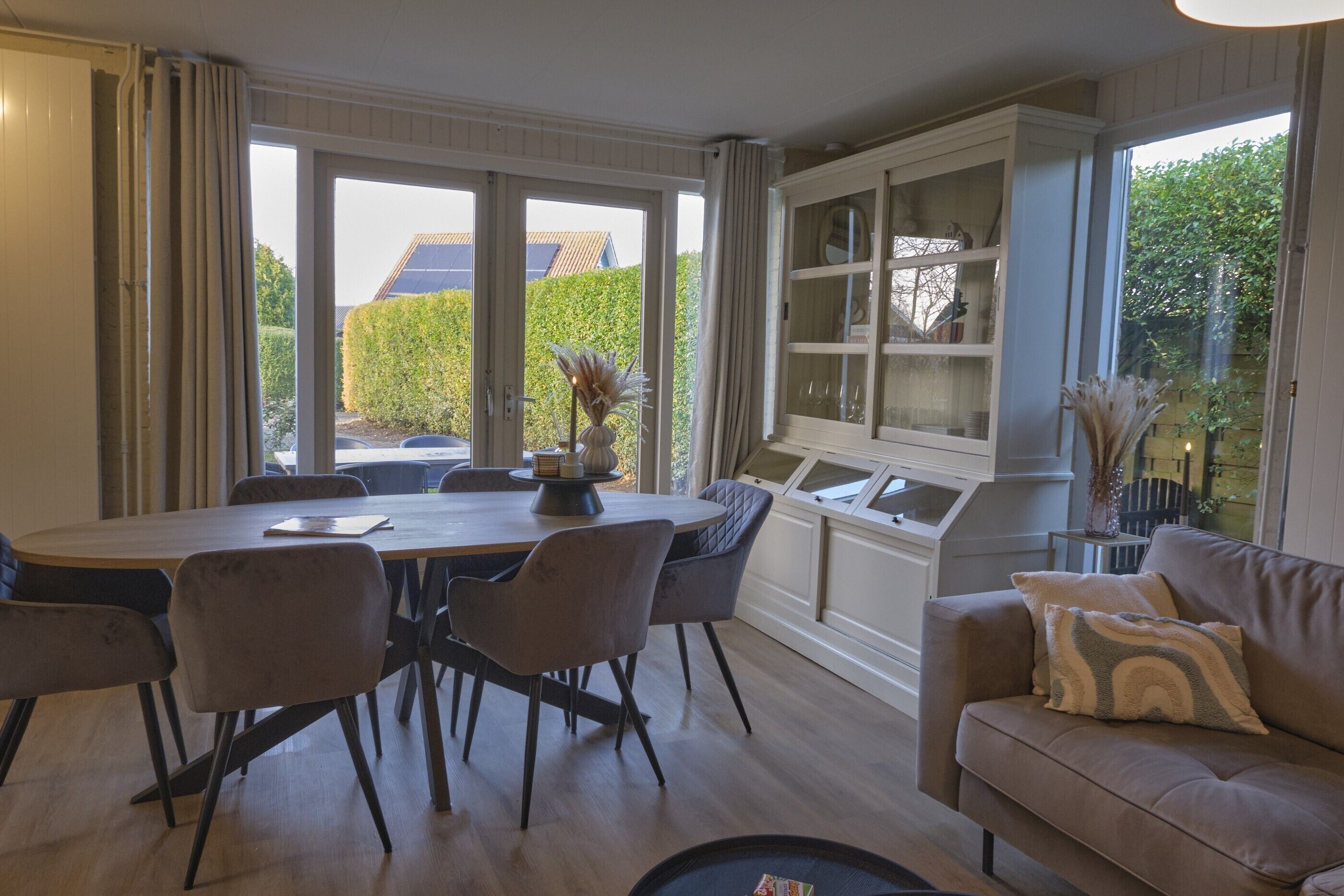 A dining area with a round table, six chairs, and a window with a view of a hedge and a house.