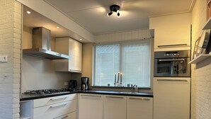 Ett kompakt kök med vita skåp, en gasspis, en kaffebryggare och ett fönster med persienner.