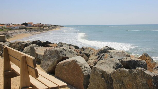 Une plage avec un banc en bois, de gros rochers et des maisons qui bordent le rivage.
