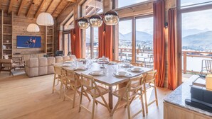 Een ruime eetruimte met een houten tafel gedekt voor een maaltijd, grote ramen met uitzicht op de bergen, en een gezellige zithoek met een televisie.