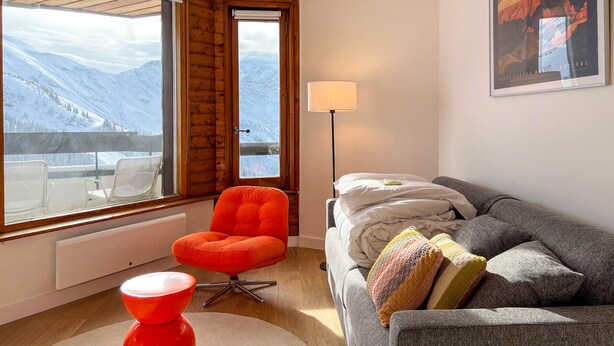 A modern room with a view of snow-covered mountains, a wooden-framed window, a bright orange chair, a grey sofa with colorful pillows, and a floor lamp. - Renovated Studio 4 Pers Avoriaz, Mountain View (Avoriaz)